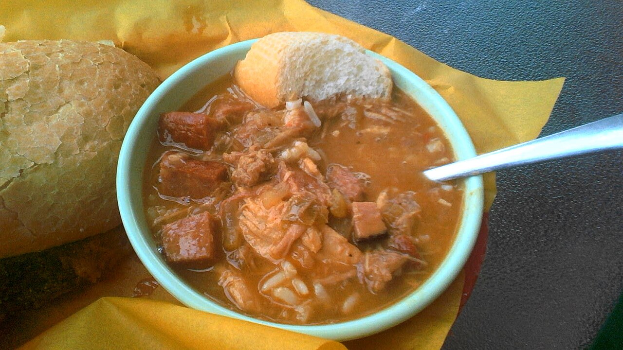 Light green bowl of gumbo with smoked sausage pieces, shredded chicken and rice in an orange-brown broth, with a crusty bread roll beside it photo_0