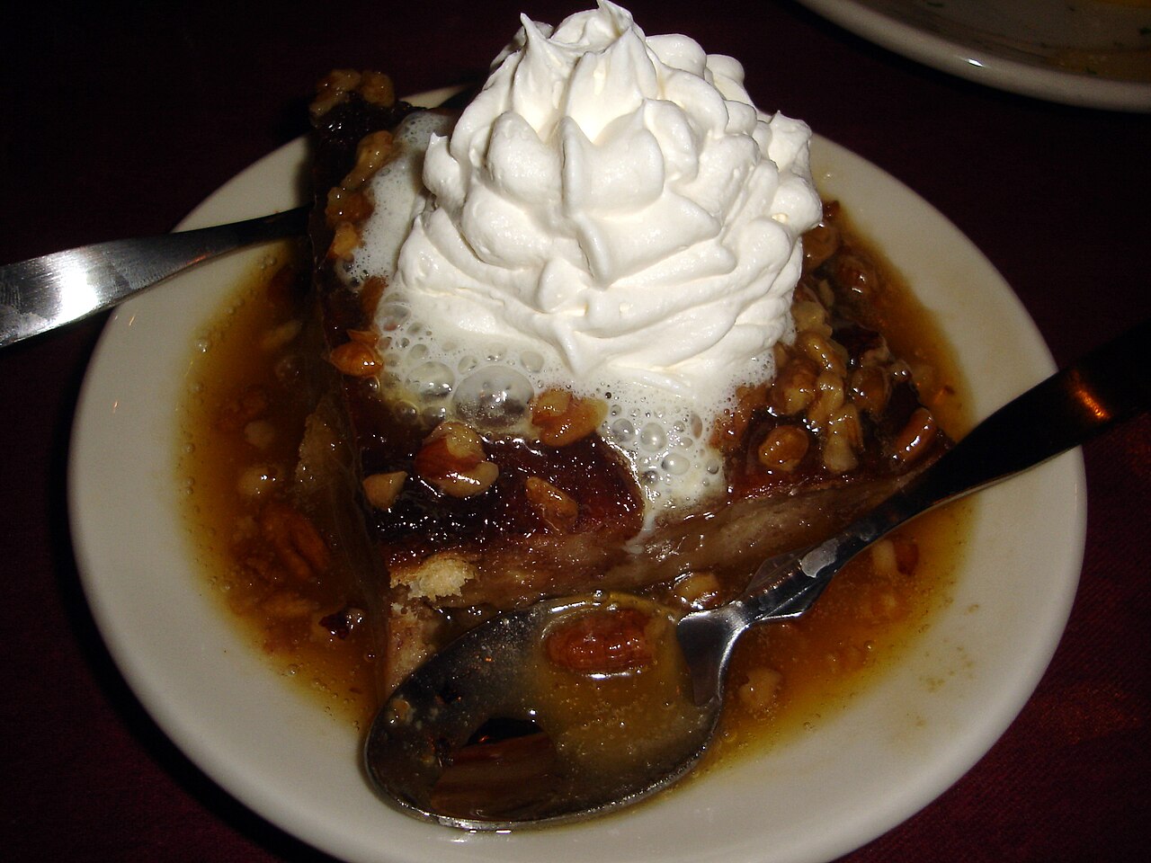 Square of bread pudding on a white plate covered in a caramel-colored pecan sauce and topped with a swirl of whipped cream photo_0